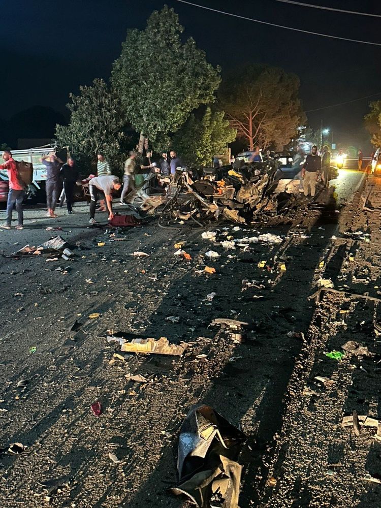A nighttime scene shows the aftermath of a drone strike on a road. The remains of a vehicle are shredded and scattered across the asphalt, with twisted metal, debris, and burnt fragments covering a long stretch of the street. Several people stand around the wreckage, some inspecting the scene, others appearing to search through debris. A few vehicles and trees line the road in the background, illuminated by vehicle headlights and street lighting. The overall scene is chaotic and clearly the result of a violent explosion.