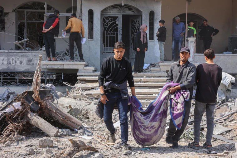 In the image, several men are seen carrying a body wrapped in a purple blanket through the rubble of a destroyed building in Gaza. The area around them is filled with debris—broken concrete, twisted metal, and splintered wood. Behind them, a group of people stands on the damaged steps of what appears to be a residential building with shattered windows and a cracked facade. The scene conveys devastation and grief, highlighting the human toll of the bombardment even amid the announcement of a ceasefire.