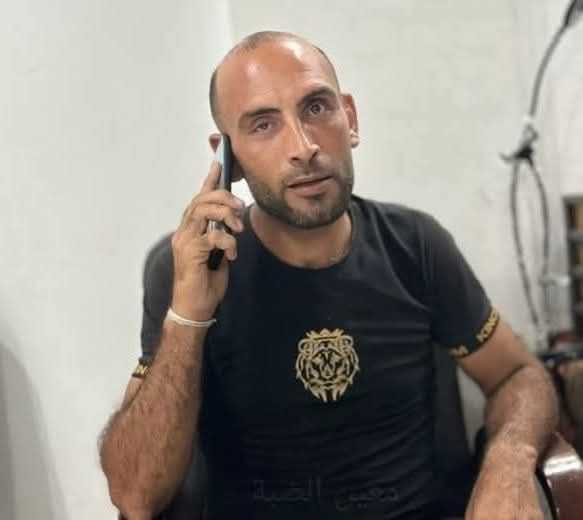 A man with a closely shaved head and trimmed beard sits indoors, holding a mobile phone to his ear. He wears a black T-shirt with a gold lion emblem on the chest and gold detailing on the sleeves. His expression is serious and focused as he looks toward the camera. The background is plain and lightly furnished, with part of a cable visible on the right.