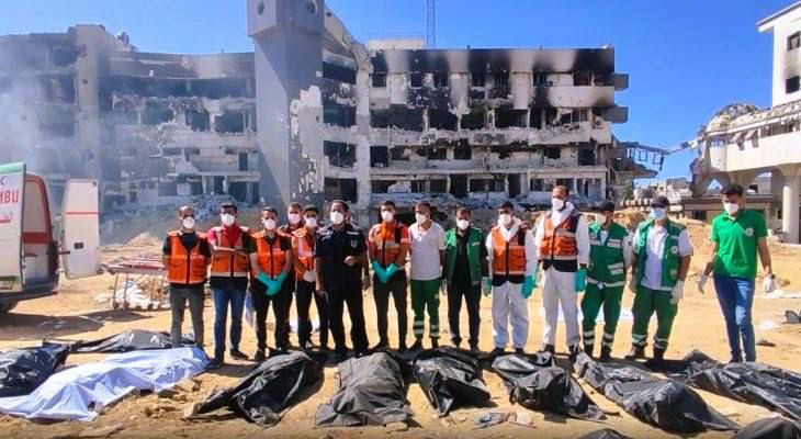 A line of Civil Defense and medical workers wearing orange, green, and white uniforms stand side by side in front of a heavily destroyed multi-story building at Al-Shifa Hospital. In front of them, numerous body bags lie on the ground, arranged in rows. Smoke and rubble surround the area under a clear blue sky.
