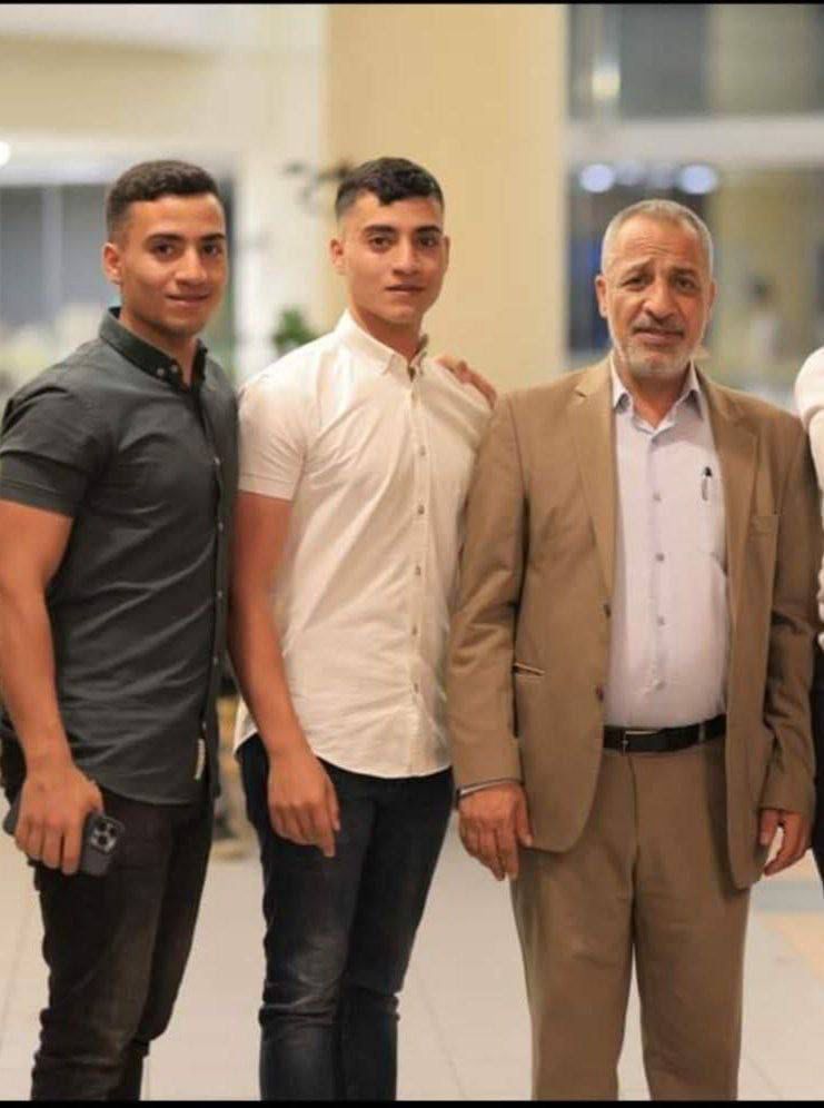 A warmly lit indoor photo shows three men standing closely together, posing for the camera. On the left are two young men with short dark hair; one wears a dark short-sleeved button-up shirt and holds a phone in his hand, while the other wears a crisp white button-up shirt with his arm resting lightly on the older man’s shoulder. The older man on the right has short grey hair and a trimmed grey beard. He wears a tan suit with a light blue shirt underneath. All three look directly at the camera with neutral to slight smiles. The background is softly blurred, suggesting a hall or reception area.