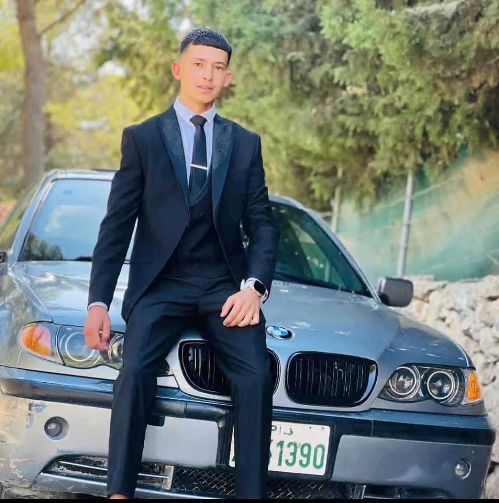 A teenage boy wearing a black suit and tie sits on the hood of a dark-colored BMW parked outdoors. He looks directly at the camera with a composed expression. Trees and a roadside area are visible in the background.