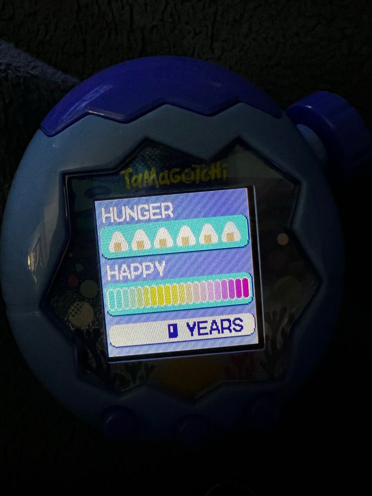 Dark image of a brightly lit tamagotchi paradise screen. There are three categories: hunger (with a full bar of onigiri beneath it), happy (with a full rainbow bar beneath it) and years (indicating zero, as in zero years). 