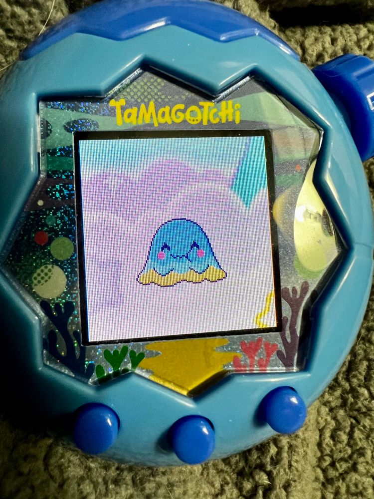 Close-up image of a blue Tamagotchi Paradise. On its screen is a little blue-and-yellow blob with rosy cheeks smiling happily. 
