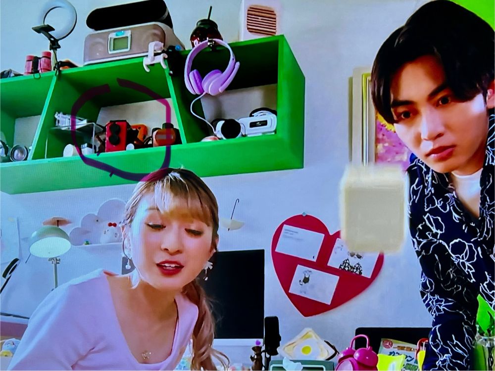 Poor quality screen shot from episode 40 of Kamen Rider Gavv. Two people, a man and a woman, are looking toward the camera as a little white box jumps in the foreground. Behind the woman is a bright green shelf and on it, highlighted with a crudely drawn circle, is a magenta camera with two lenses.