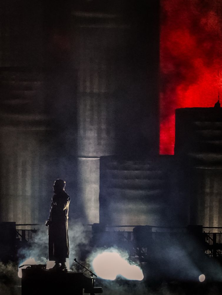 Image taken from the grass area of the amphitheater. Tyler is in his trench coat and standing on his piano. Behind him is Dema. You see red behind the towers - looks kind of like a fire is burning l. 