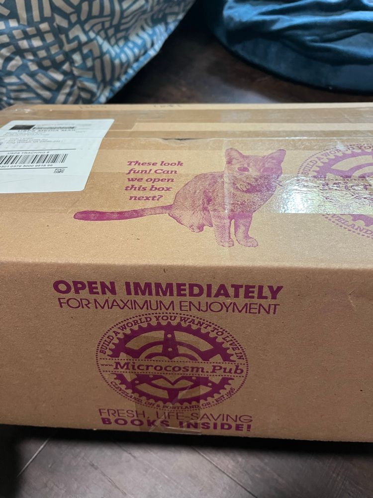 A brown cardboard box with purple ink printed on it. On the top flap, it has a kitty cat looking at you saying “these look fun! Can we open the box next?”. On the side of the box: it says “open immediately for maximum enjoyment”. Then, there’s a microcosm gear logo. Under that it says “fresh, life – saving books inside!”. The logo reads “build a world you want to live in. Cleveland Ohio and Portland Oregon. Established 1996