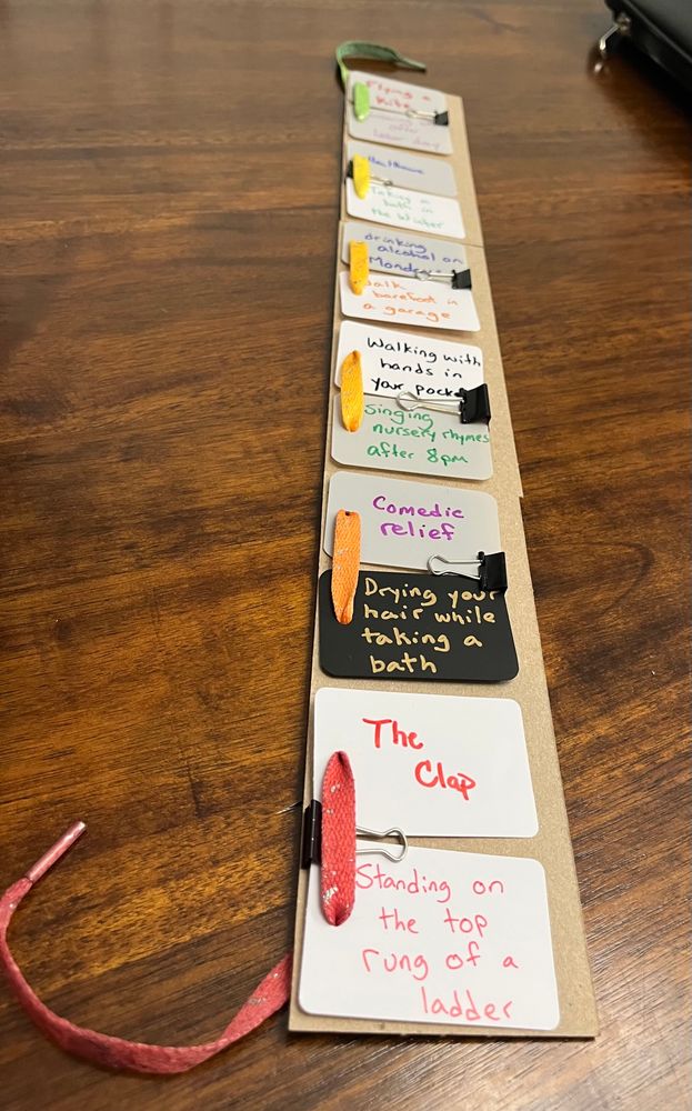 The bottom cards read: “comedic relief”, “drying your hair in the bath”, the clap”, “standing on the top rung of the ladder”. A long skinny board just wider than applying card has multiple sets of half poker cards clipped together and then along the edge of the long skinny board there’s a shoelace woven in one card and out another an in one and the other in one and out the other all the way down, the shoelace goes from green at the top to red at the bottom