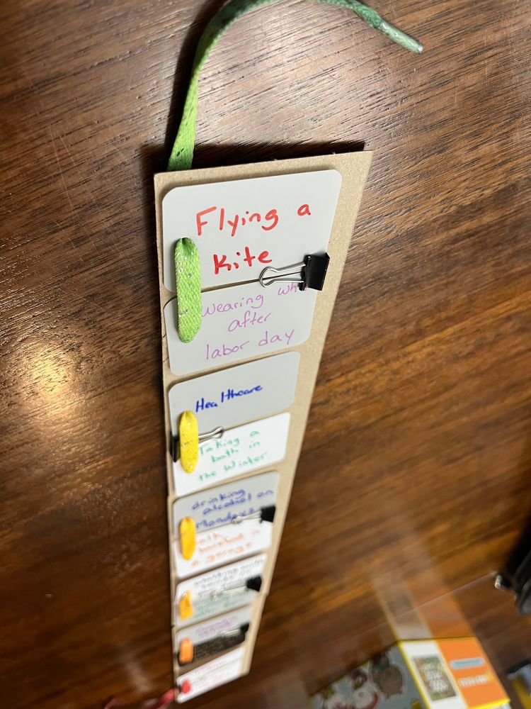 Top cards read: “flying a kite”, “wearing white after Labor Day”, and “healthcare”. A long skinny board just wider than applying card has multiple sets of half poker cards clipped together and then along the edge of the long skinny board there’s a shoelace woven in one card and out another an in one and the other in one and out the other all the way down, the shoelace goes from green at the top to red at the bottom