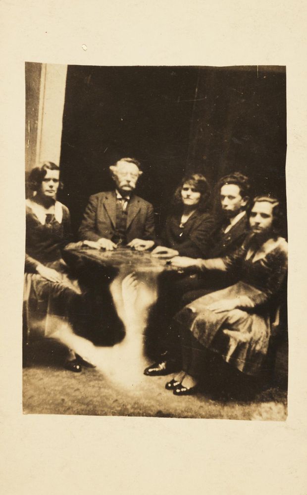A black and white photograph of a séance. A hand rises from the mist pooling near the floor. 