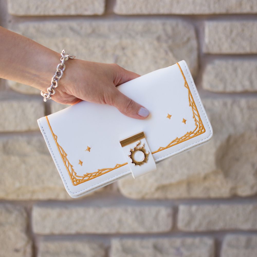 A white clutch style wallet with the meteor symbol from FF14. There is gold embroidery to mimic the menu style in the game. It is being held by the model's hand for size comparison.