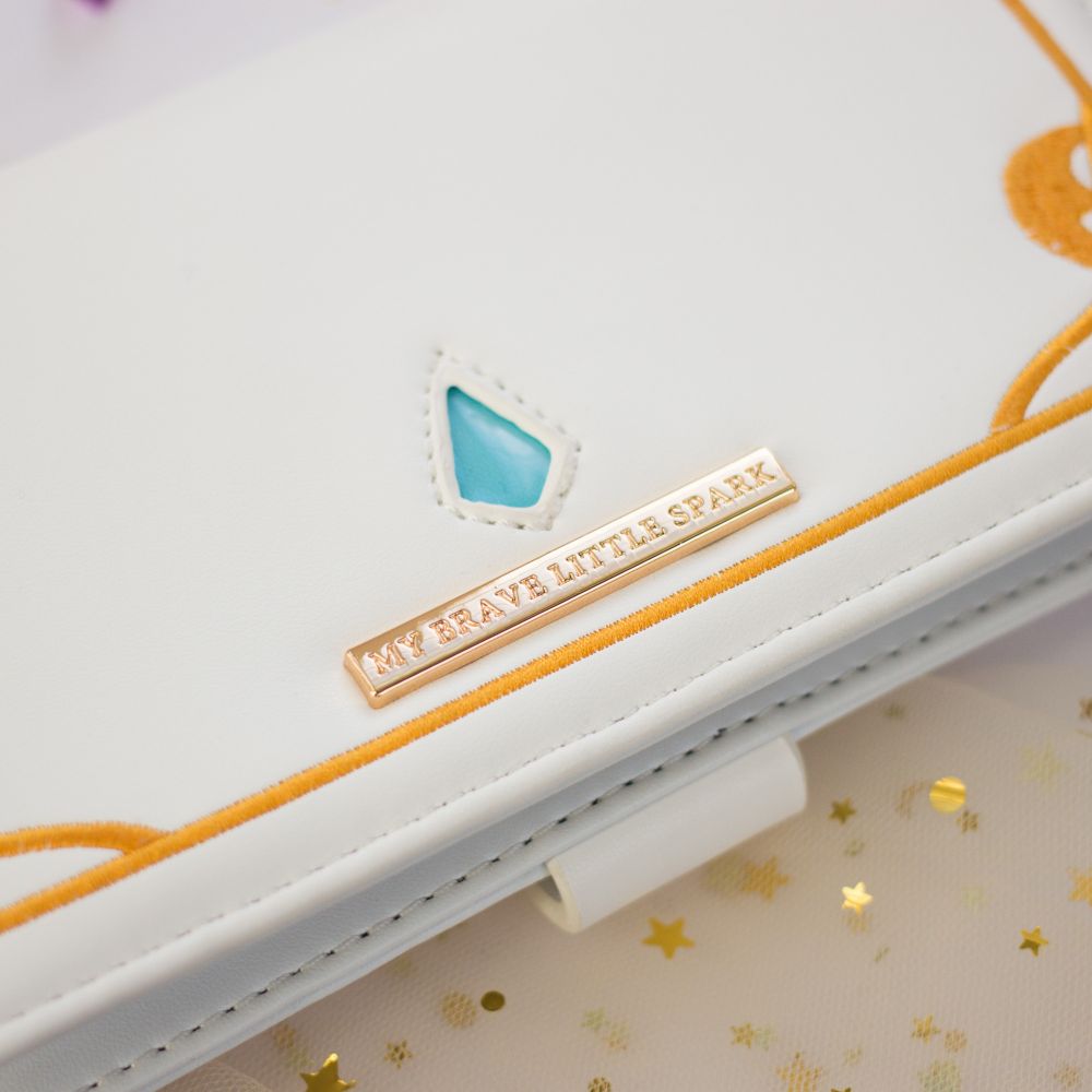 A white clutch style wallet with a cut out of the crystal hydaelyn and a gold hardware plaque that is engraved saying "MY BRAVE LITTLE SPARK".
