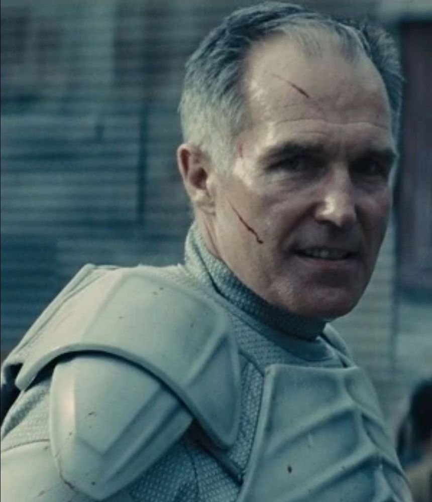 A menacing soldier with body scratches on his face, dressed in Stormtrooper white body armor over white spandex uniform, standing in front of corrugated metal walls.