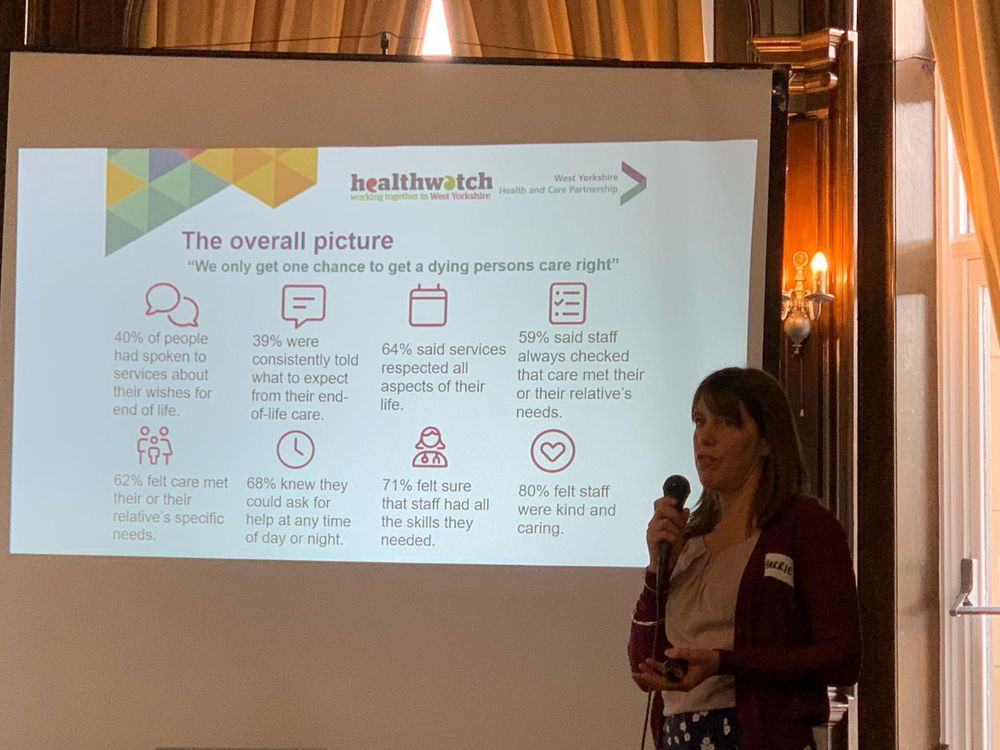 A person with long hair speaking into a microphone with a presentation screen behind that shows some statistics about end of life care