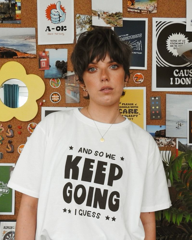 Jenny is a slim white woman with short dark hair, wearing a white tshirt with black text that reads ‘and so we keep going I guess’.