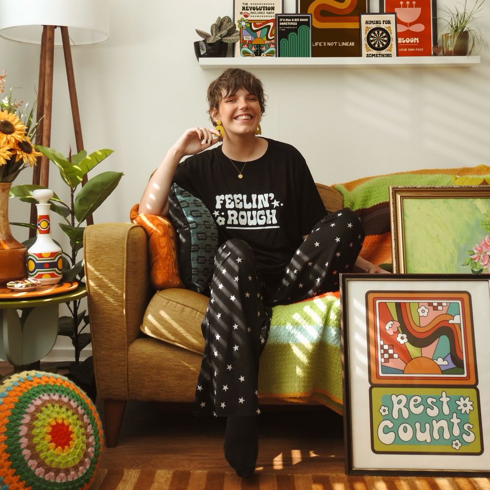 Jenny is a white slim woman with short dark hair sitting on a sofa smiling. She is wearing a black tshirt with white text on the chest that reads ‘feelin rough’.