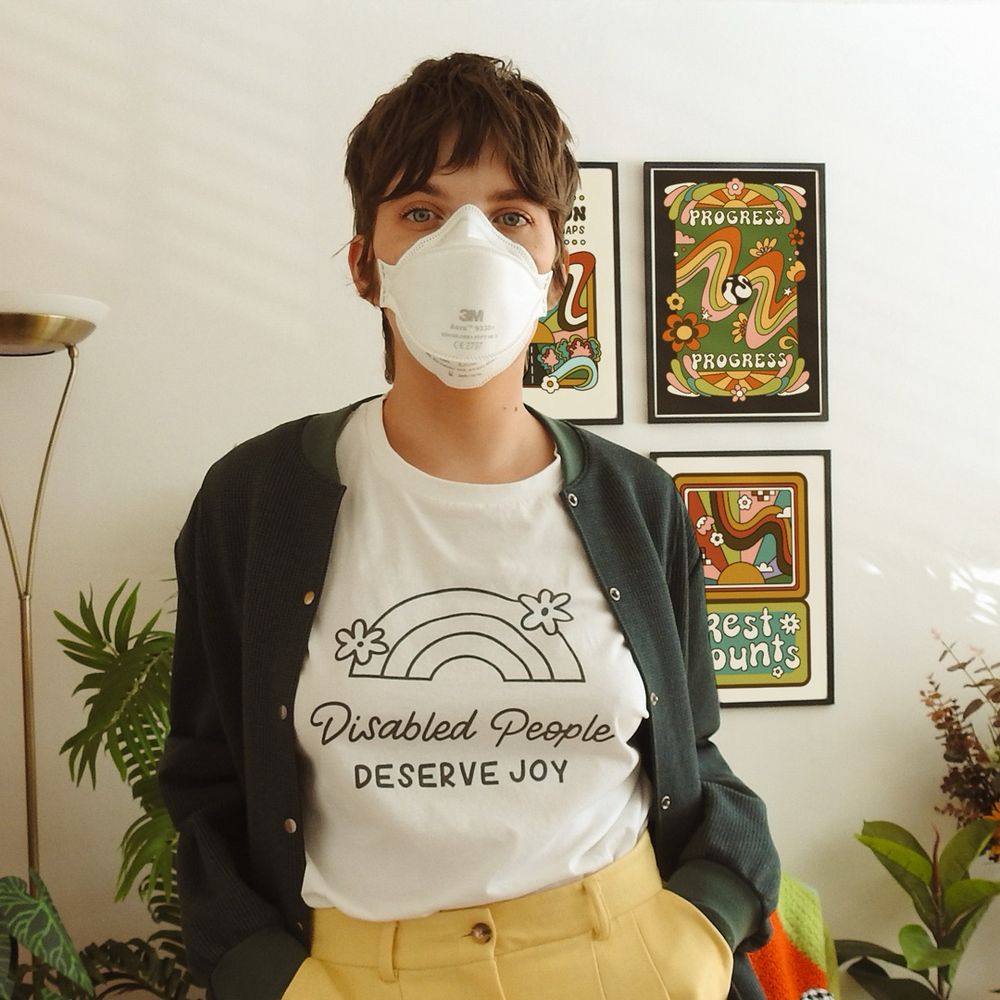 Jenny wears a white respirator mask and a white tshirt that reads ‘disabled people deserve joy’