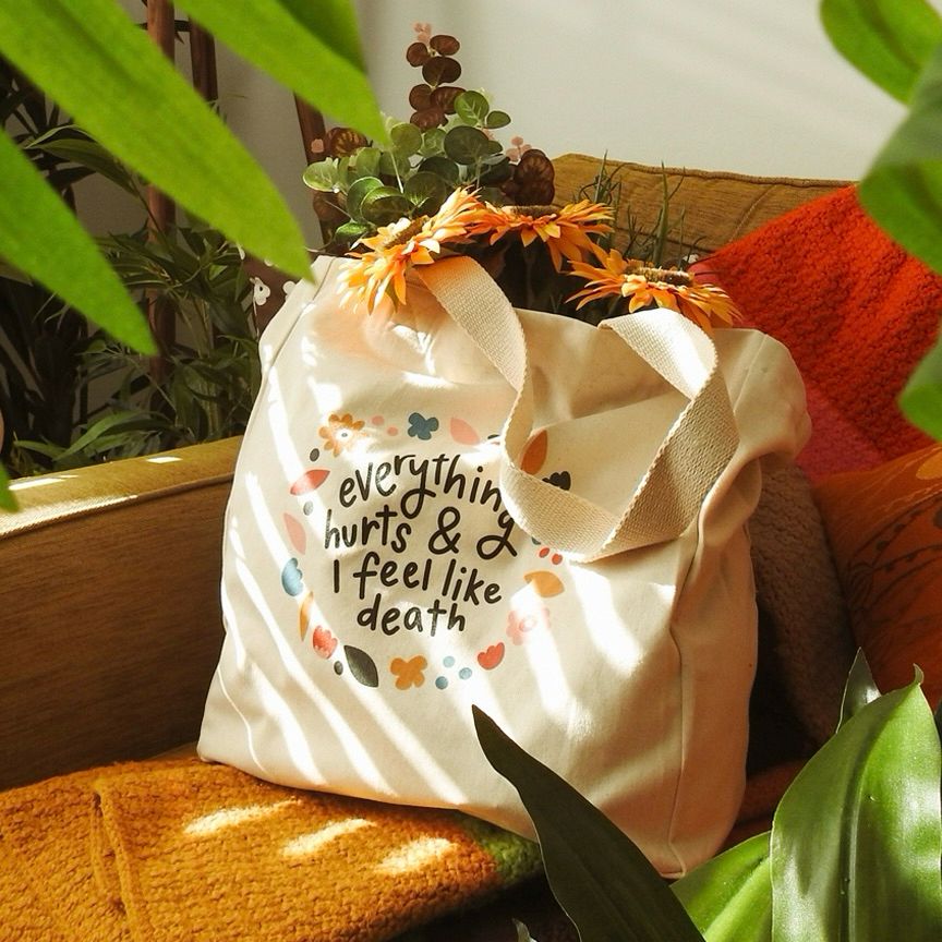 Canvas tote bag sitting on a sofa. On the front of the bag is handwritten text that reads ‘everything hurts and I feel like death’. The text is framed by colourful illustrated floral shapes.