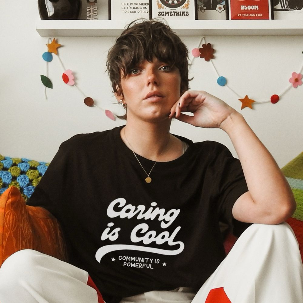 Jenny, a white slim woman with short dark hair is sitting on the sofa at home. She’s wearing a black tshirt with white text on the chest that reads ‘caring is cool, community is powerful’