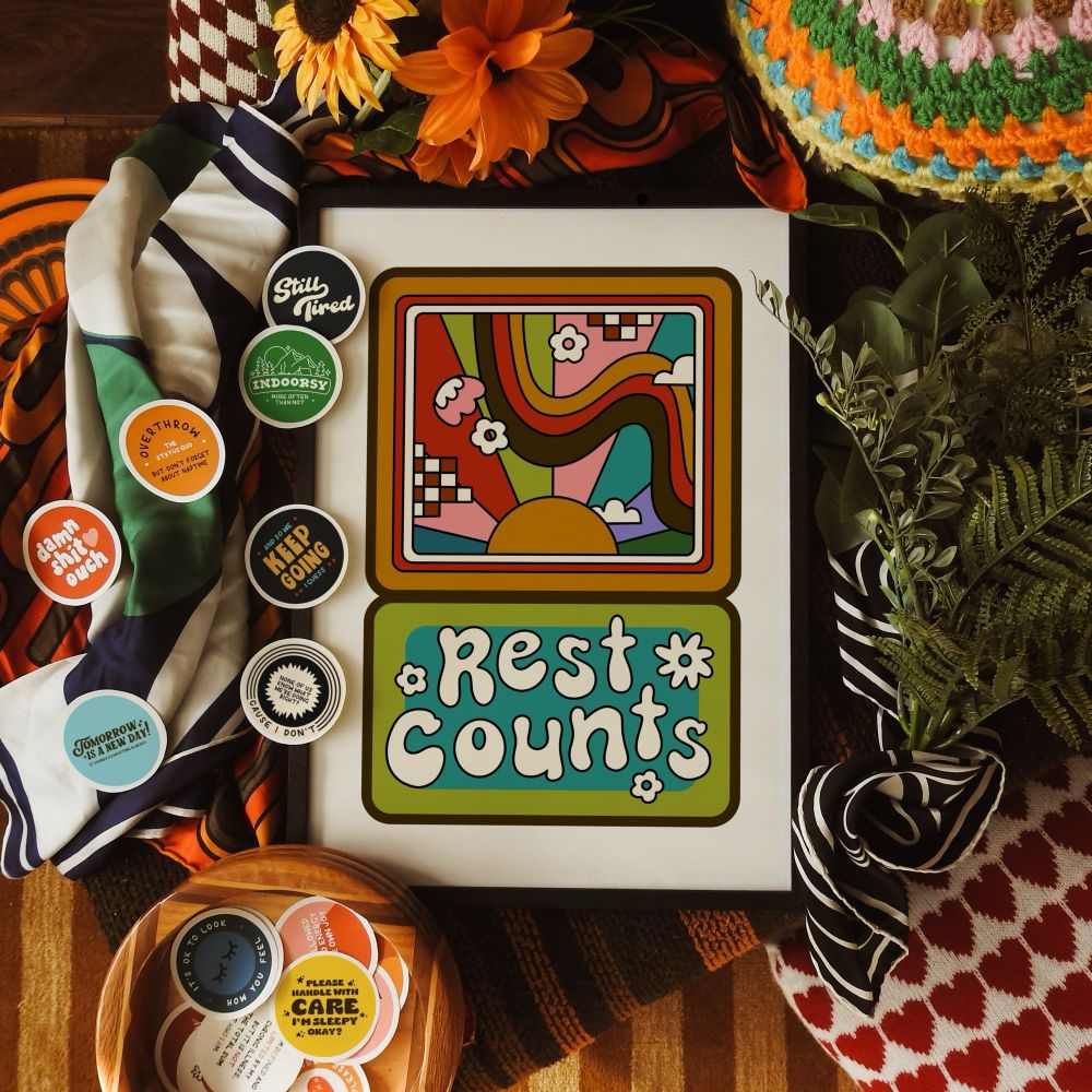 Framed art print displayed against colourful patterned fabrics. Print reads ‘rest counts’, and features a retro illustration of sunbeams and florals.