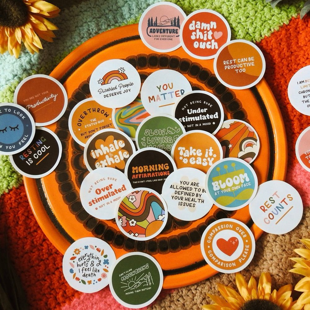 Selection of round stickers displayed on a colourful blanket. Stickers feature phrases such as ‘bloom at your own pace’, ‘take it easy’, and ‘resting is cool’