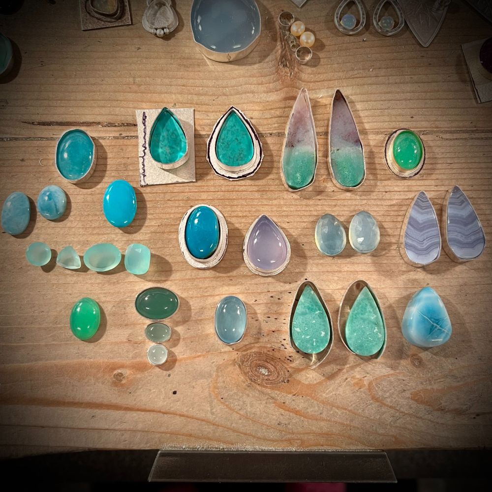 A view of a variety of gemstones arranged on my wooden jeweller's bench.  The stones are in summery colours of aqua, green, turquoise, blue and lavender.
