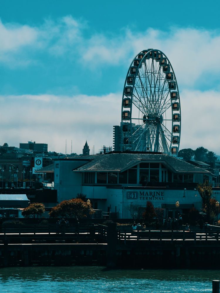 A large Ferris wheel rises behind the historic Pier 41 Marine Terminal along San Francisco’s waterfront under a partly cloudy sky.