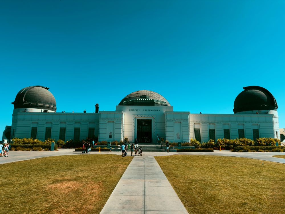 A front view of Griffith Observatory in Los Angeles, with people walking around the entrance on a clear day.
