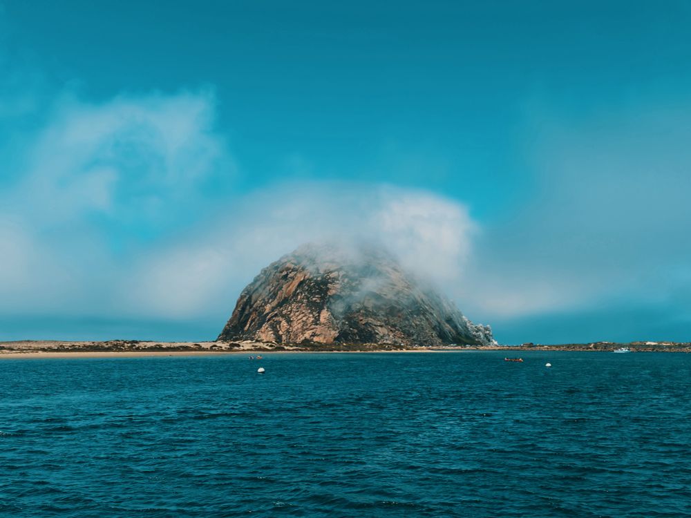 Morro Bay Rock, a rocky volcanic plug covered in sparse foliage, protruding from the waters of Morro Bay, California.