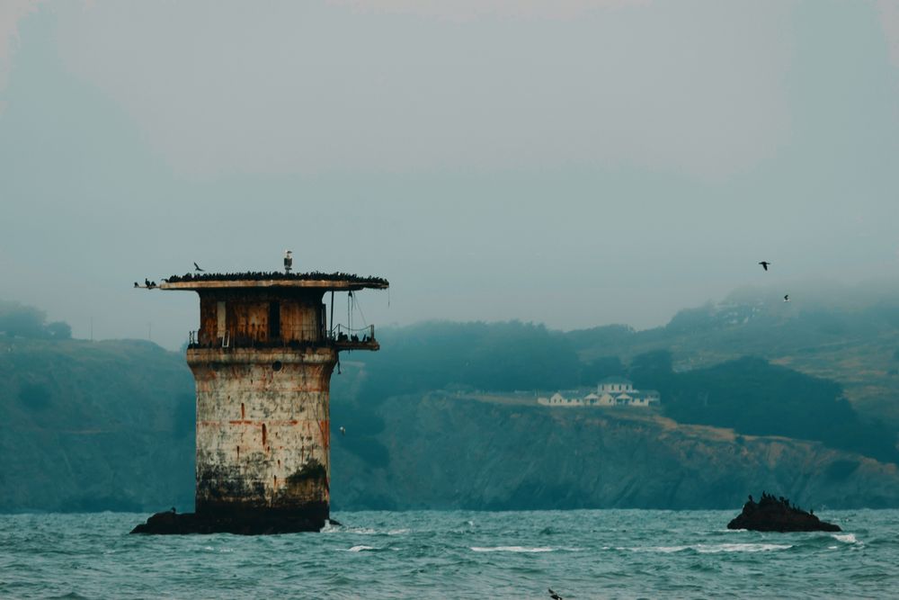 An old, rust-stained lighthouse stands on a rock in the ocean with foggy hills and buildings in the background.