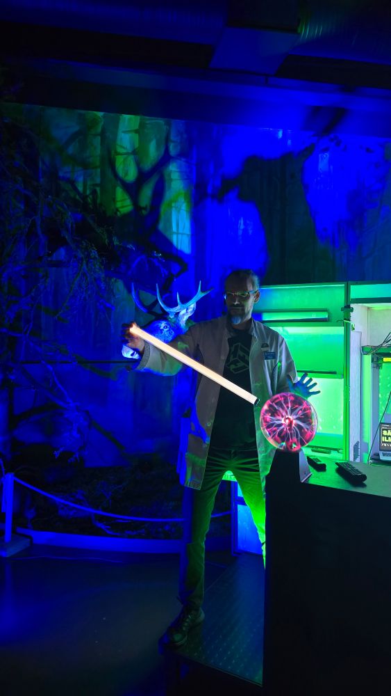 Värivalojen valaisema Kristjan sytyttää loisteputken plasmapallon avulla. Olan yli kurkkii täytetty hirvi.