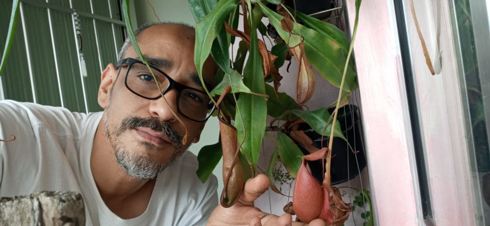 A foto é retangula, atras uma porta de aluminio, na frente eu , branco, cabelo, barba e bigodes curtos e grisalhos, estou olhando pra camera, segurando um jarro de nepenthes, planta carnivora.