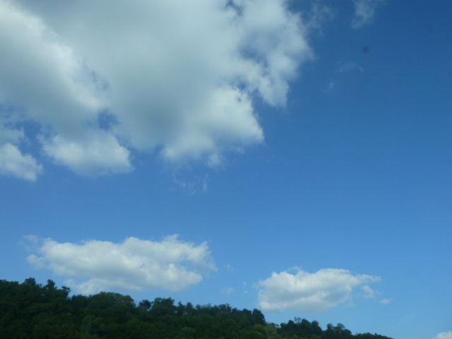 Big, white clouds with blue sky over green trees on the top of a hill.