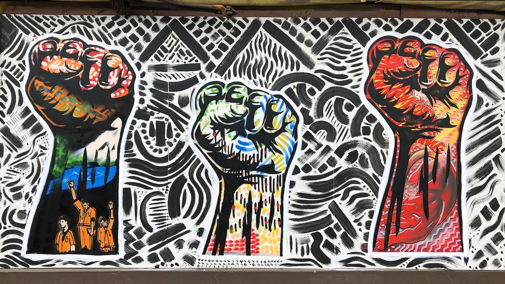 A photo of a mural graffiti featuring three colorful raised fists, the symbol of solidarity. One of the fists has a depiction of the 1968 Olympics Black Power salute incident during which two African-American athletes, Tommie Smith and John Carlos, each raised a black-gloved fist during the playing of the US national anthem.

The background of the mural features various geometrical shapes in black on a white background.