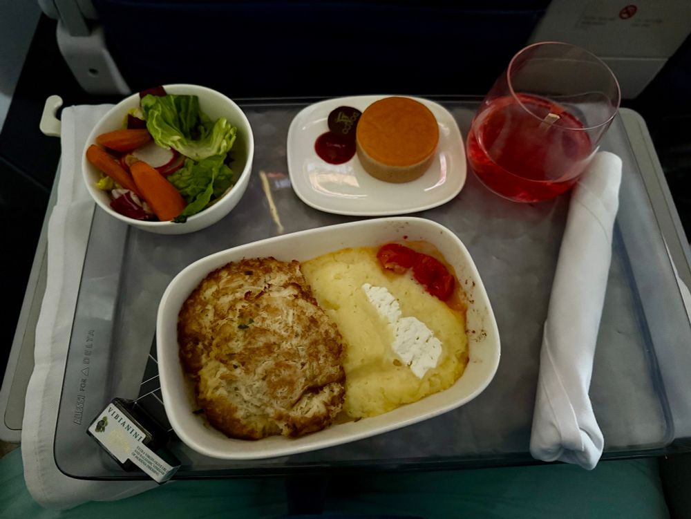 Airline meal on a tray table featuring a main dish of crab cakes and mashed potatoes topped with sour cream and cherry tomatoes. Sides include a small salad with lettuce, carrots, and radishes, a mini tart-style dessert, a glass of pink beverage, and a wrapped napkin with utensils.