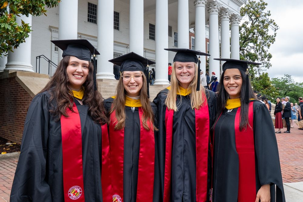Four graduate students pose in regalia in front of the Reckord Armory