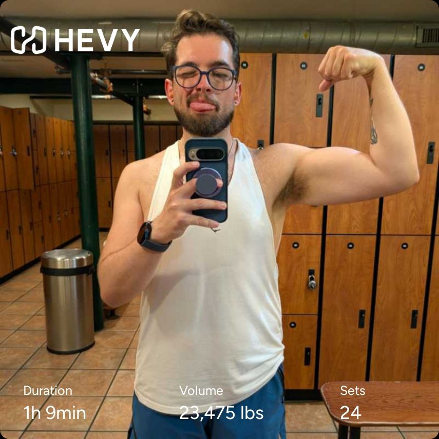 Gym locker room mirror selfie of me in a white tank top and teal shorts flexing my right bicep and sticking out my tongue. Hevy workout info included 