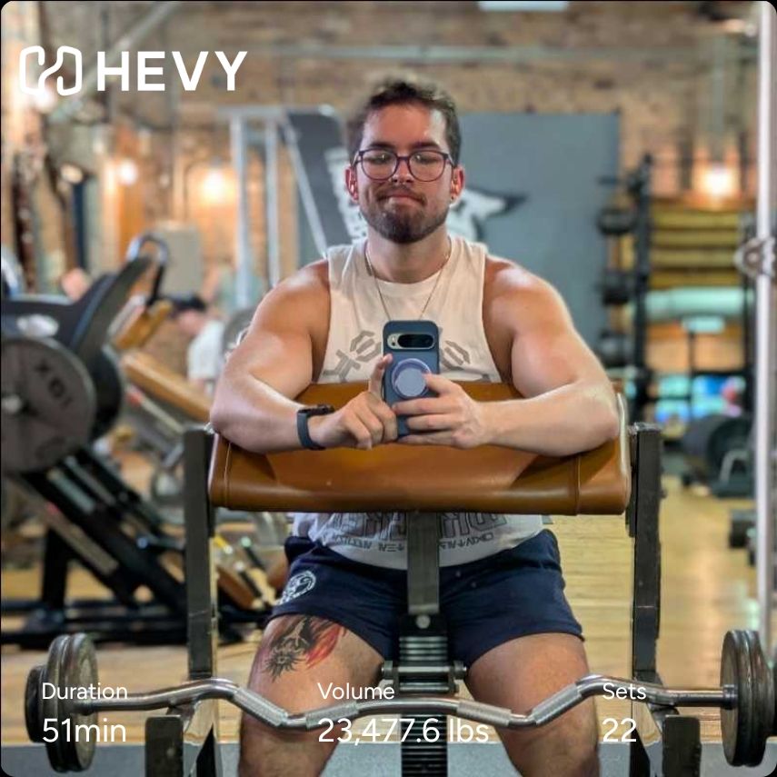 Gym mirror selfie of me at the preacher curl stand in a white cut off shirt and navy shorts. Hevy workout info included. 