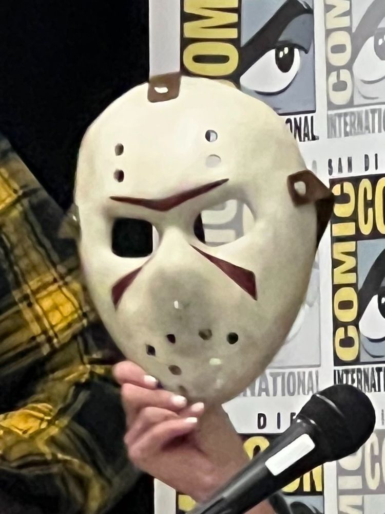 Screenshot from the stream of a panel at San Diego Comic-Con 2025: a hand holds up a newly designed Jason Voorhees hockey mask during announcements of upcoming Friday The 13th media.