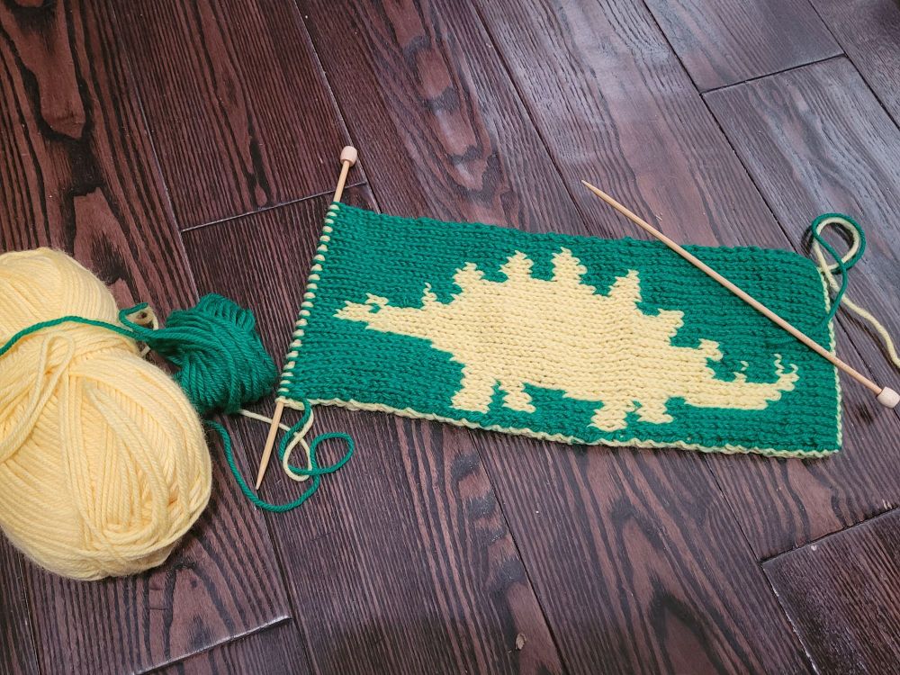 Reverse side of the same double-knitted scarf, showing a yellow stegosaurus on a green background. 