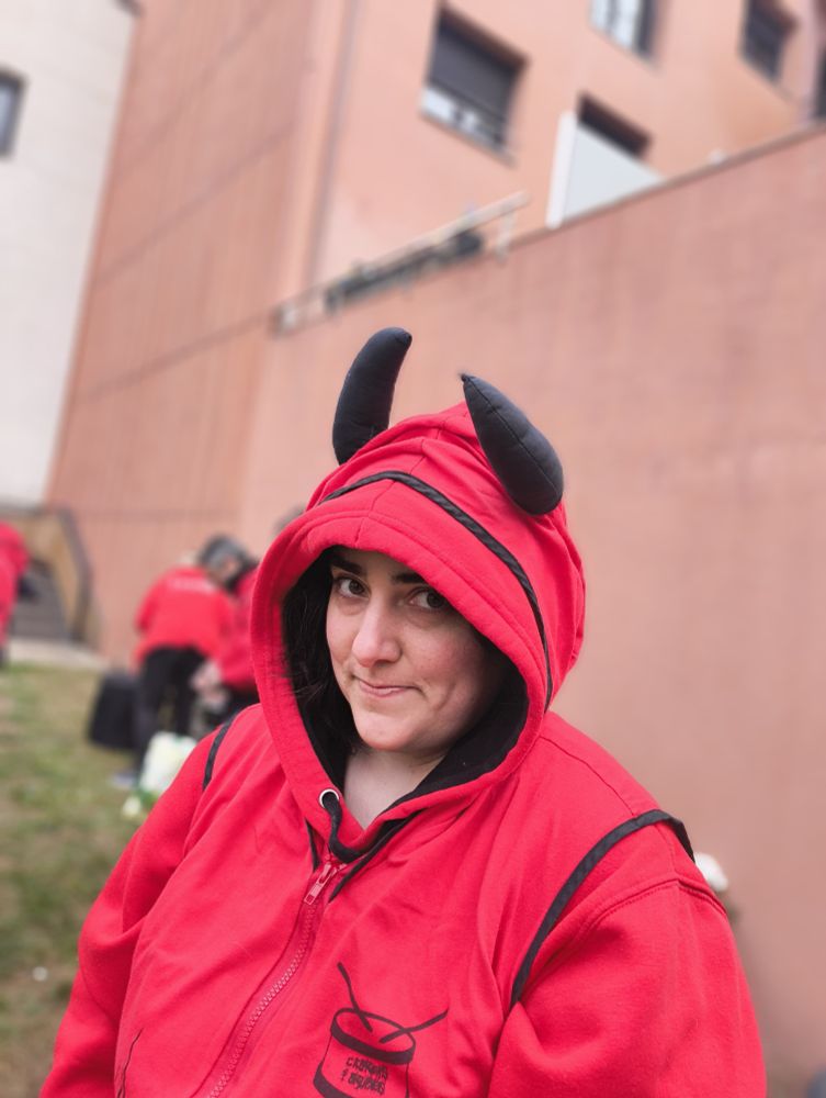 Un miro de ladrillos en el fondo y hierba en el suelo desenfocado, hay dos personas al fondo y una chica en el centro mirando a la camara con una sudadera roja y un chaleco rojo por encima con un logo de un tambor en su pecho izquierdo. Lleva la capucha puesta, que lleva dos cuernos de tela negra.