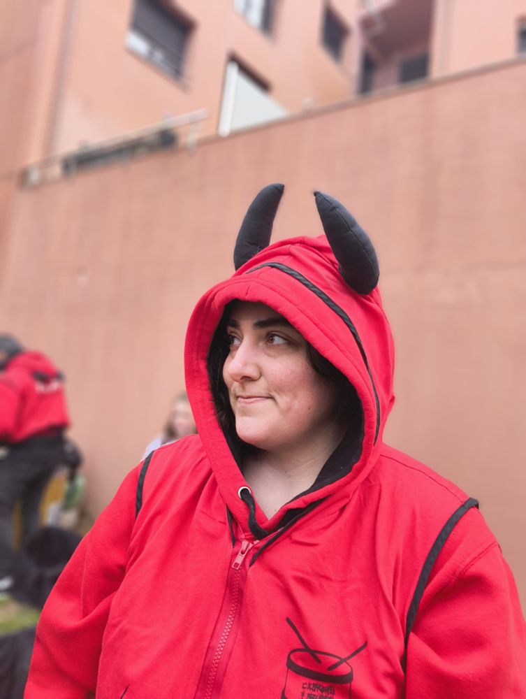 Un miro de ladrillos en el fondo y hierba en el suelo desenfocado, una chica en el centro mirando al lado con una sudadera roja. Lleva la capucha puesta, que lleva dos cuernos de tela negra.