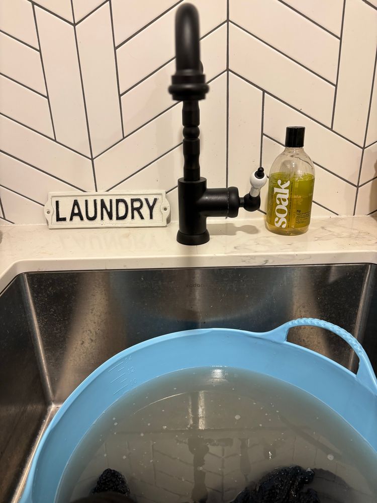White tiled laundry room and sink with blue laundry basin.  Grey cashmere scarf soaks in the basin with Soak laundry soap. The water in the basin is grey, dirty. 