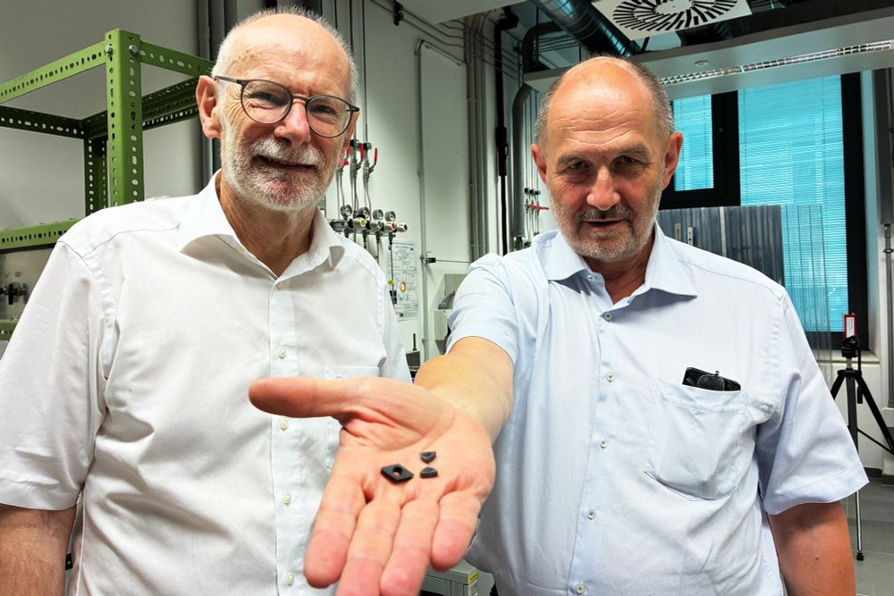 Zwei Männer stehen im Labor, einer streckt seine Hand nach vorne, auf der schwarze Muttern liegen.