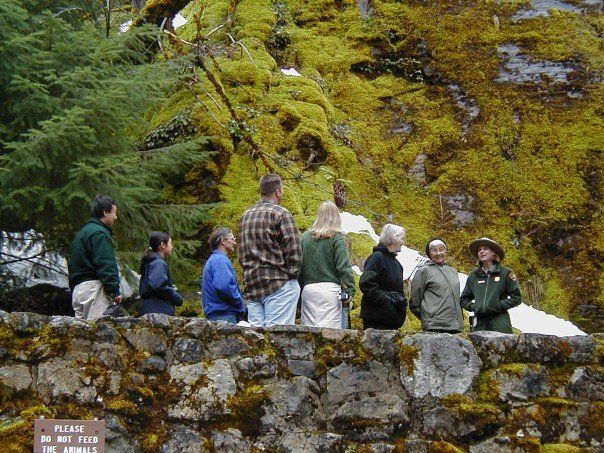 A group of people standing in front of a moss covered rock face. At the right a woman in a park service uniform stands facing the group. 