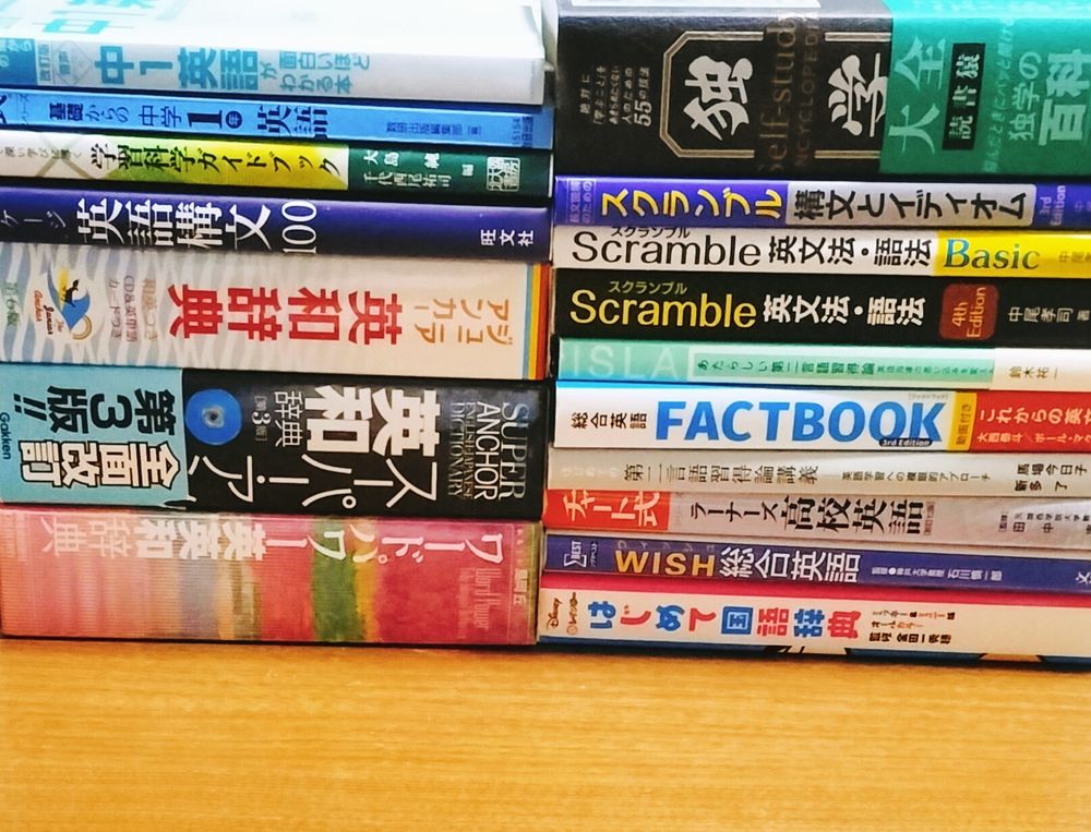 ​分厚い英語辞書や文法書、理論書が2列に積み上げられた写真。
​【左の山：辞書・中学英語】
上から「中1英語が面白いほどわかる本」「基礎からの中学1年英語」「学習科学ガイドブック」「ステージ 英語構文100」「ジュニア・アンカー英和辞典」「スーパー・アンカー英和辞典 第3版」「ワードパワー英英和辞典」。
​【右の山：文法・独学・SLA】
上から「独学大全（読書猿）」「スクランブル構文とイディオム」「Scramble 英文法・語法 Basic」「Scramble 英文法・語法 4th Edition」「あたらしい第二言語習得論の講義」「総合英語 FACTBOOK」「外国語学習の科学 第二言語習得論講義」「チャート式 ラーナーズ高校英語」「WISH総合英語」「ディズニー はじめて国語辞典」。
​初心者向けから専門的な第二言語習得論（SLA）、大学受験用の網羅系参考書まで、幅広いレベルとジャンルの書籍が揃っている様子。