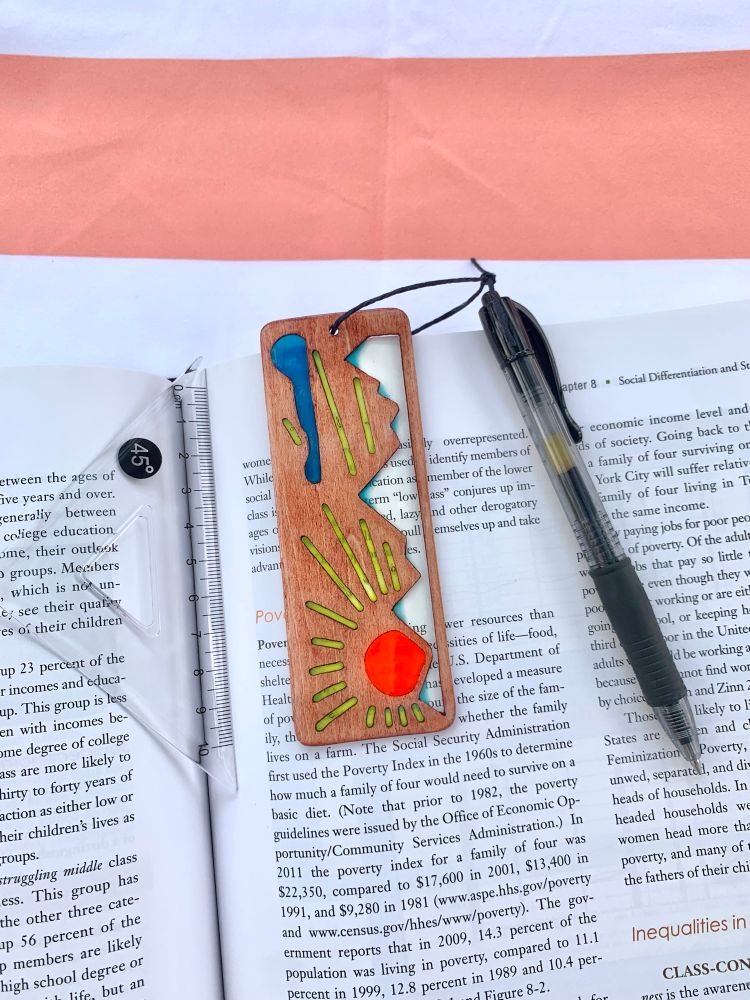 Mountain sunrise wood and resin bookmark laying on an open textbook next to a pen and a 45 degree triangle ruler for size.
