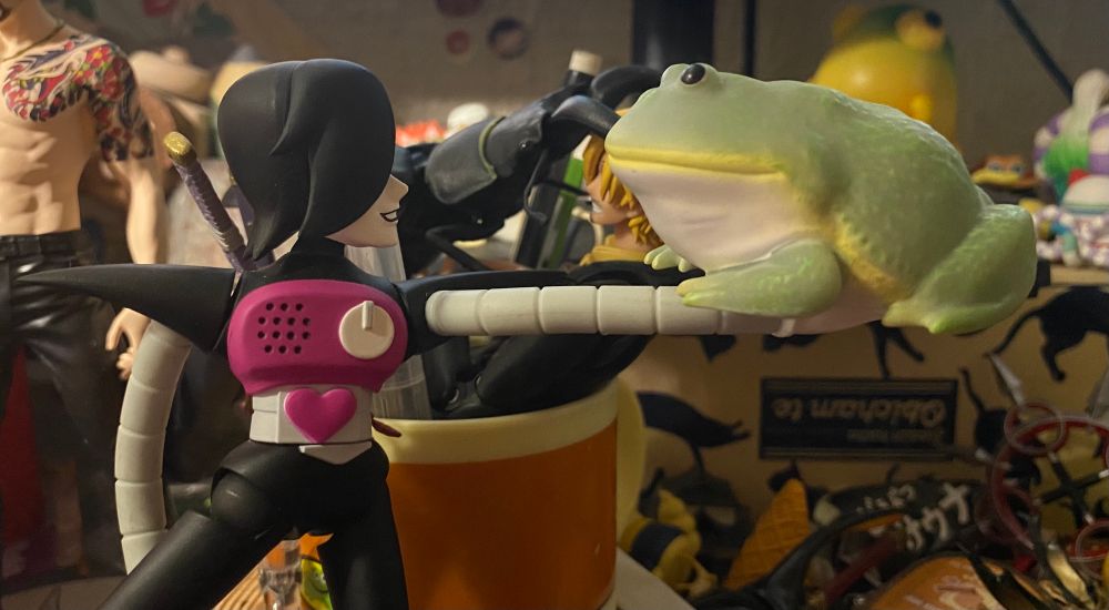 Mettaton figure holding a big green frog in his hand, smiling at it. Behind him, Sanji is being attacked by a giant beetle, but this ain’t about him. Majima’s there too 