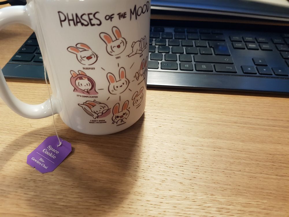 A photo of my desk in the office, on it a keyboard and a tall white mug. On the mug there's drawings of a comic bunny, titled "phases of the mood" displaying it sleepy, happy, and more. Out of the mug there hangs the tag of a tea bag, indicating the flavor "Space Cookie", an organic spiced chai by the manufacturer "samova".
