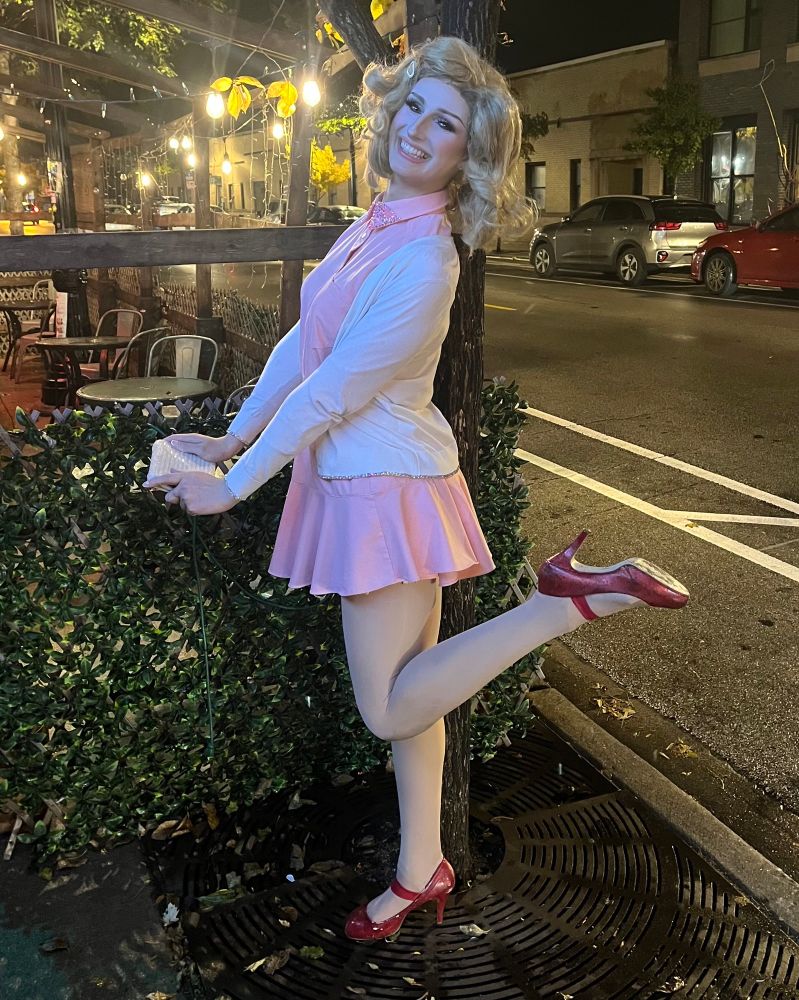 me dressed as janette, a spoof character of janet from rocky horror, posed with my leg popped up behind me and my clutch extended low away from me